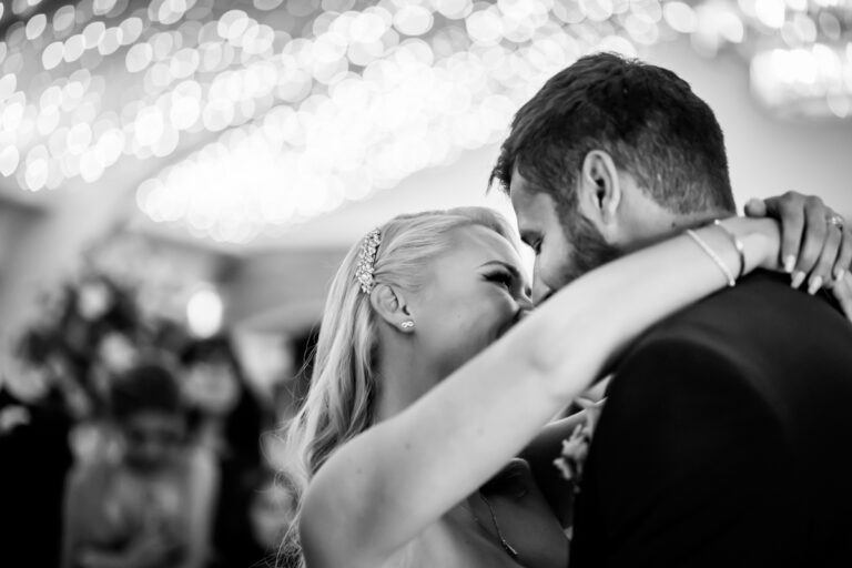 A black-and-white photo of a couple embracing and smiling at their wedding. The bride has long blonde hair adorned with a headband and the groom has short hair and a beard. In the background, out-of-focus guests smile and twinkling lights create a glowing ambiance. Image by Andy Wade Photography.