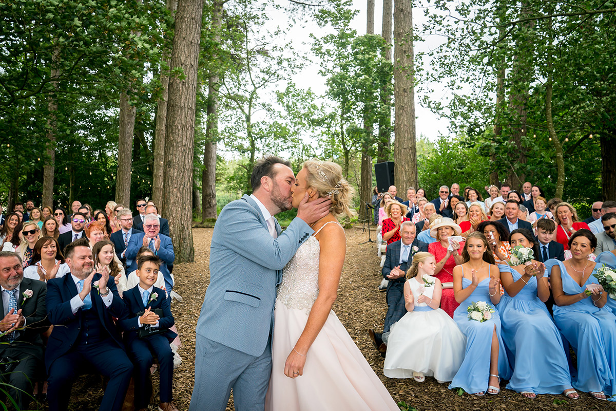 A bride and groom share a kiss at their outdoor wedding ceremony in Lancashire, surrounded by tall trees. The bride wears a white gown and the groom a light blue suit. Guests seated on either side, including bridesmaids in light blue dresses, applaud and smile at this picturesque wedding venue. Image by Andy Wade Photography.
