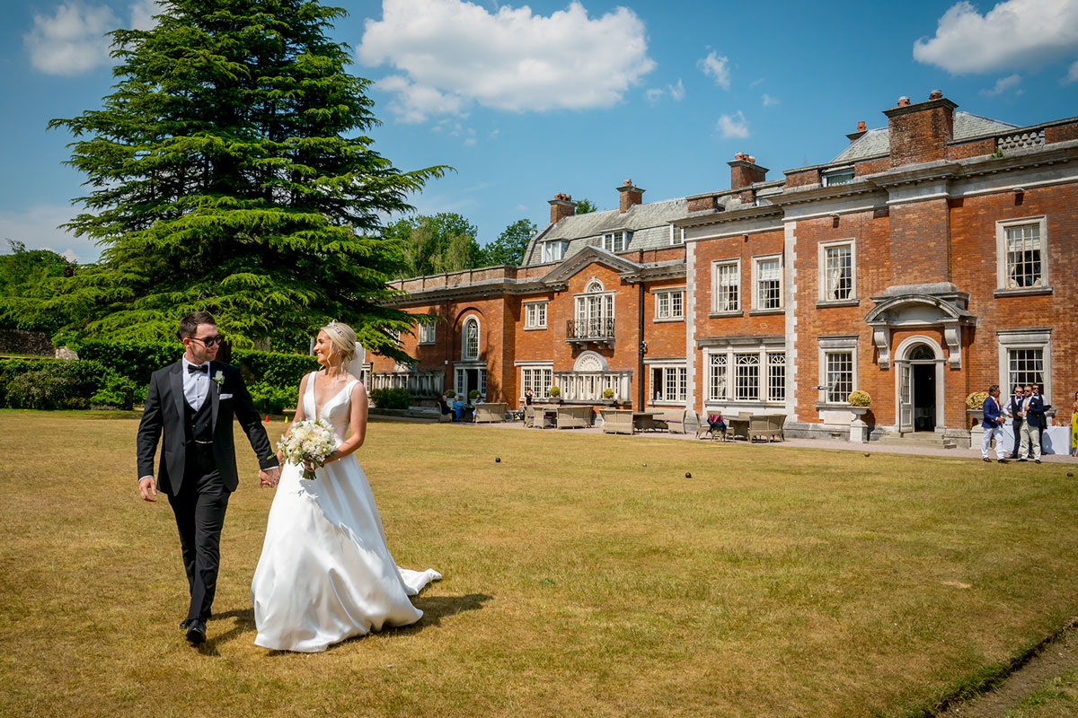 A bride and groom stroll hand-in-hand across the lawn of an elegant manor house with red brick walls and numerous windows. The bride is in a white gown holding a bouquet, and the groom is in a black tuxedo. A large tree and a blue sky with clouds are in the background. Image by Andy Wade Photography.
