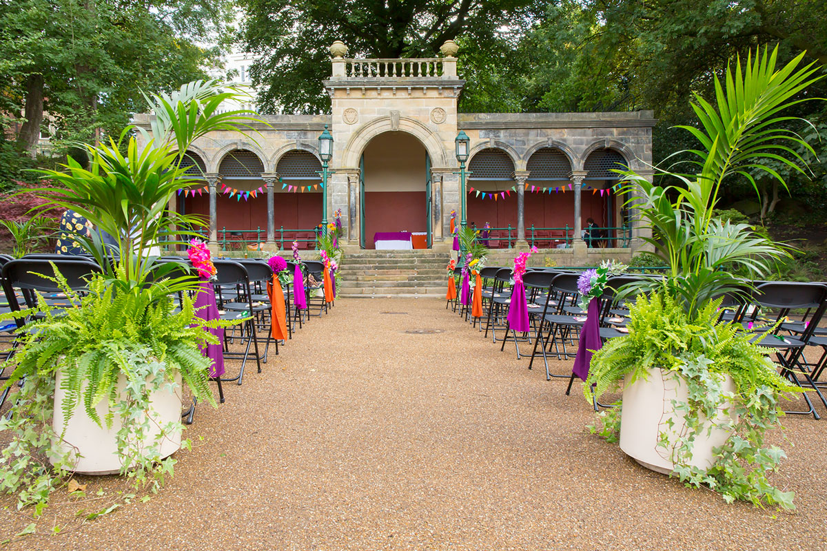 A beautifully decorated outdoor wedding setup with rows of chairs adorned with colorful ribbons, vibrant plants along the aisle, and a historic stone pavilion in the background. The pavilion is festively adorned with multicolored pennant flags. Image by Andy Wade Photography.