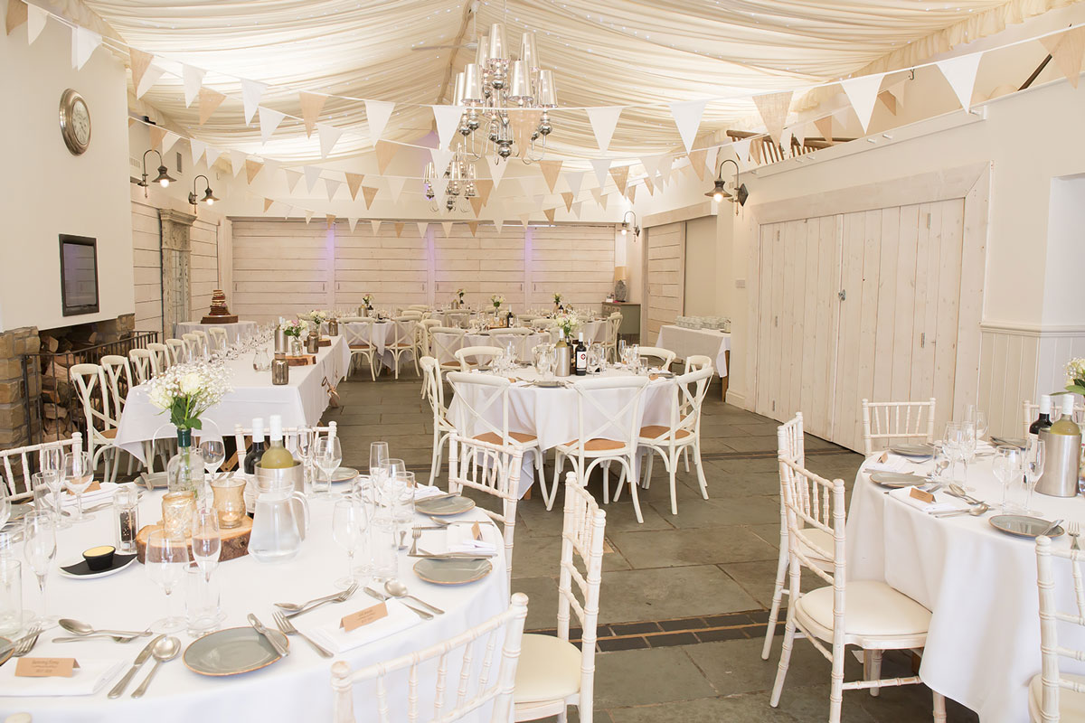 A wedding reception hall in one of the finest wedding venues in Lancashire is decorated with white and beige bunting and fairy lights. Large round tables adorned with white tablecloths, elegant tableware, flowers, and candles are set up. Twinkling chandeliers hang from the draped ceiling. Image by Andy Wade Photography.