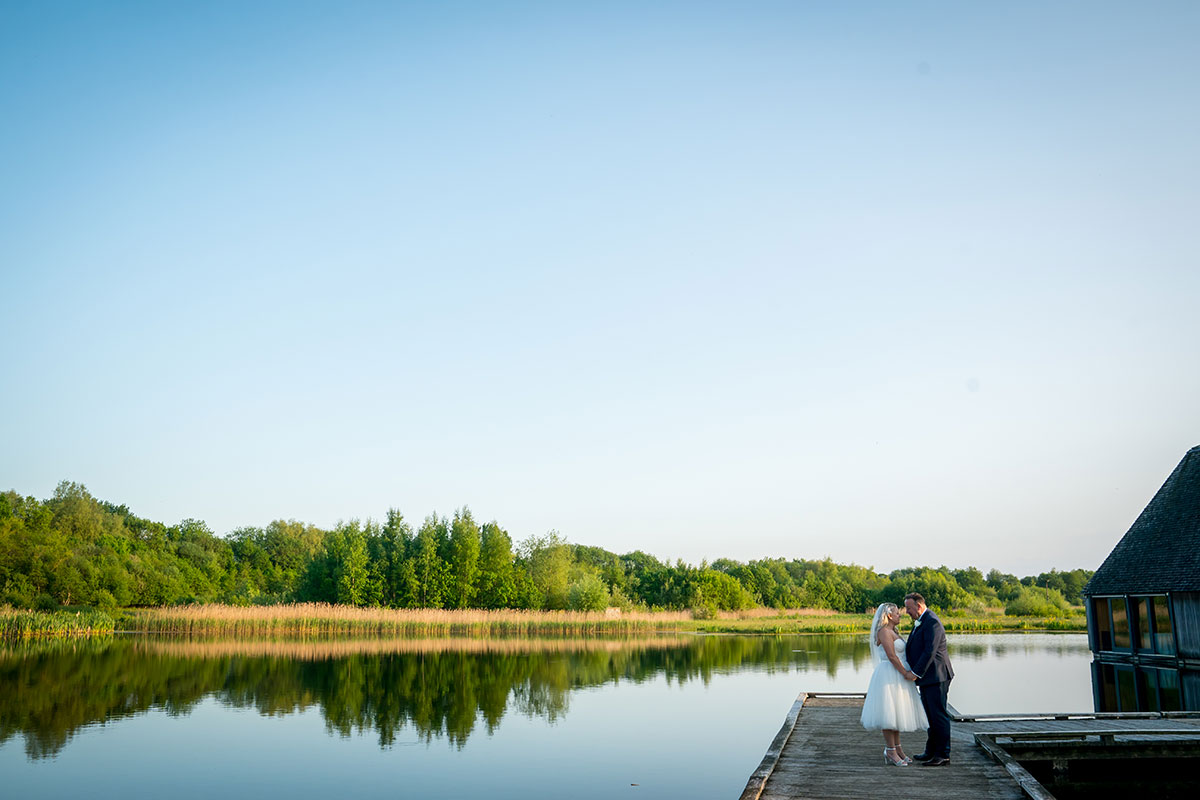 A couple dressed in wedding attire stands on a wooden pier by a calm, reflective lake in one of Lancashire's charming wedding venues, surrounded by lush green trees under a clear blue sky. The bride holds a bouquet, and the groom stands beside her, both facing each other. Image by Andy Wade Photography.