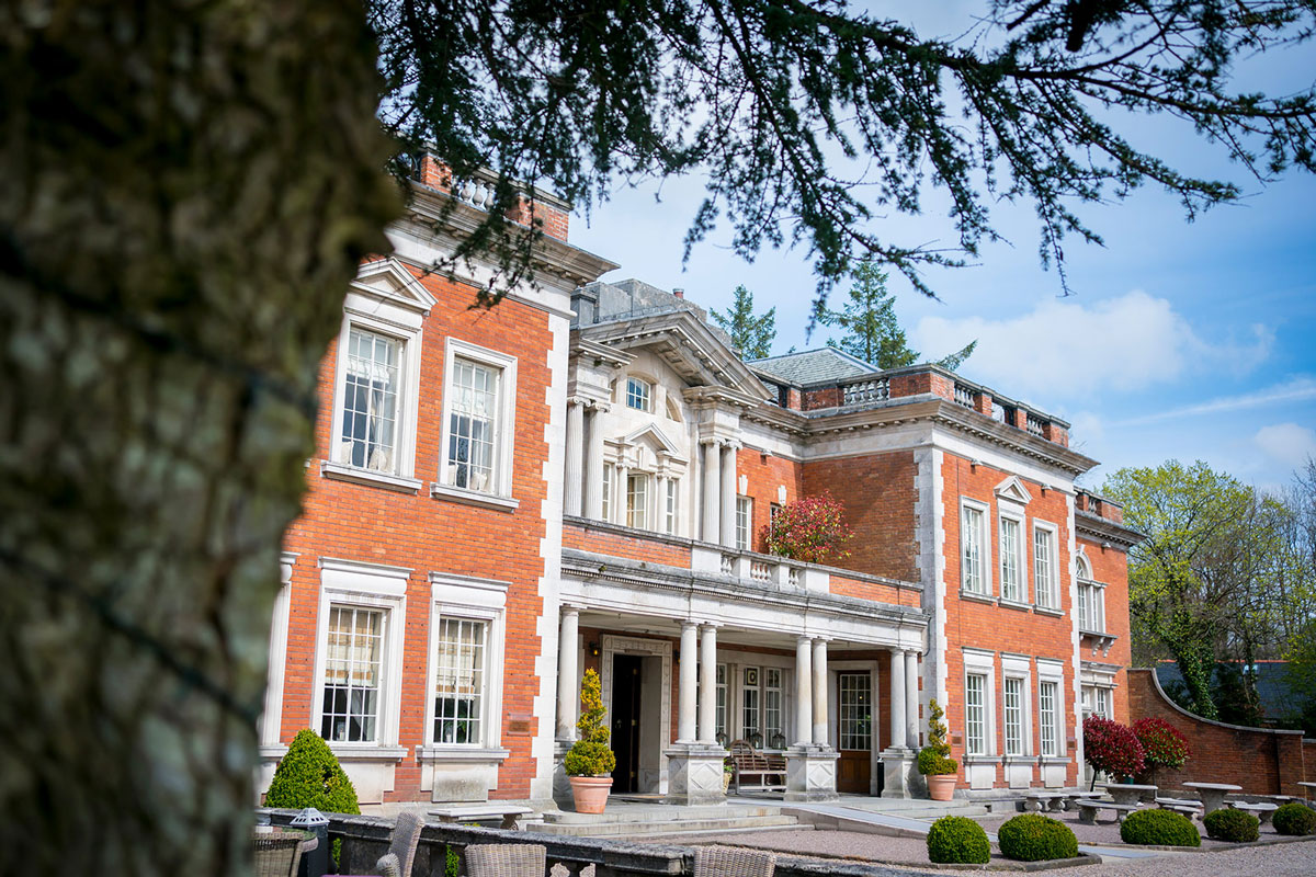 A stately red-brick mansion with white stone accents under a clear blue sky. The building features large windows, decorative columns at the entryway, and is surrounded by trees and manicured shrubs. The photo is taken with a tree in the foreground. Image by Andy Wade Photography.
