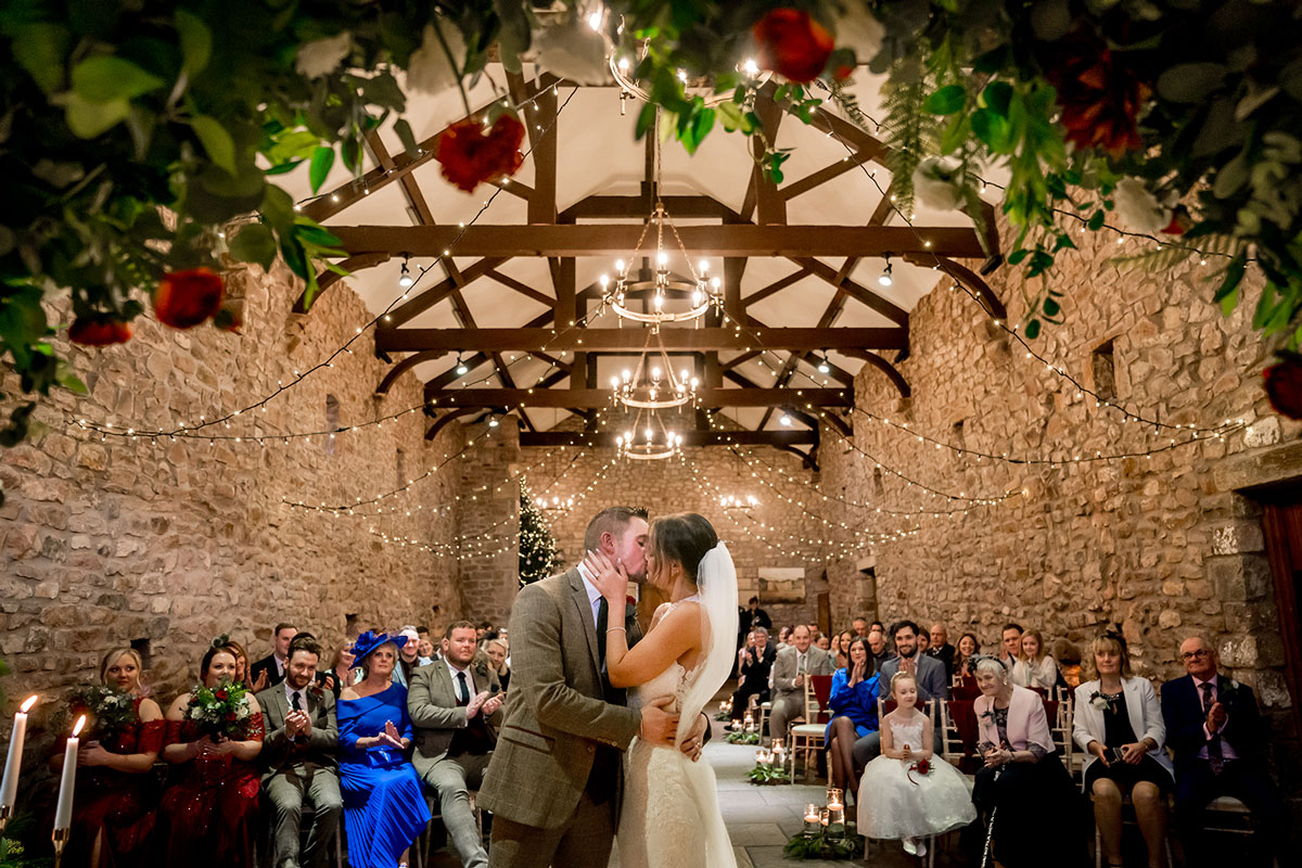 A bride and groom share a kiss at the altar of a rustic, stone-walled Lancashire wedding venue adorned with greenery and string lights. Guests are seated on either side, with some taking photos. A flower girl and other attendants in formal attire are present. Image by Andy Wade Photography.