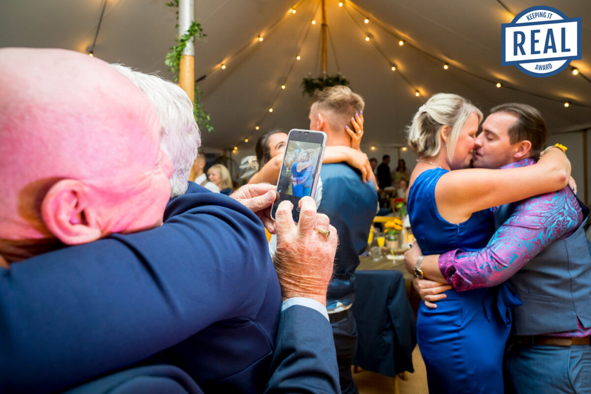 A joyful wedding celebration under a tent with string lights. Two couples are embracing and kissing. An older man with a phone is taking a photo of one of the couples. Guests in the background are socializing. A "Keeping it Real" award logo is on the top right. Image by Andy Wade Photography.