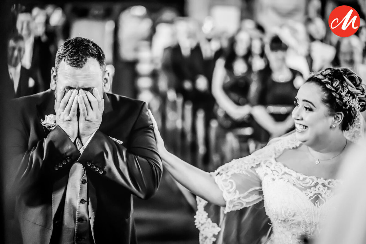 A black and white photo captures an emotional wedding moment. The groom stands with his hands covering his face, overwhelmed with emotion, while the bride, wearing a lace gown, smiles comfortingly, with her hand on his shoulder. Onlookers are visible in the background. Image by Andy Wade Photography.