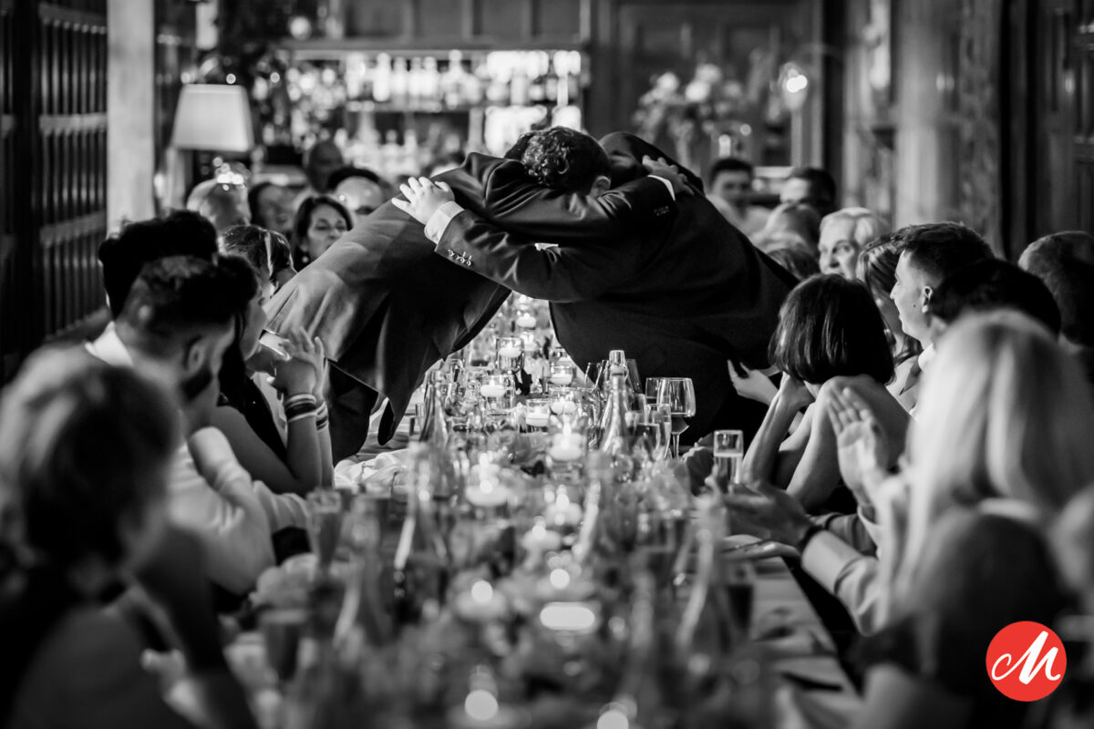 A black-and-white photo of two men hugging over a long, elegant dinner table. The table is filled with glasses and plates, and surrounded by guests who are watching and applauding. The atmosphere is warm and celebratory. Image by Andy Wade Photography.