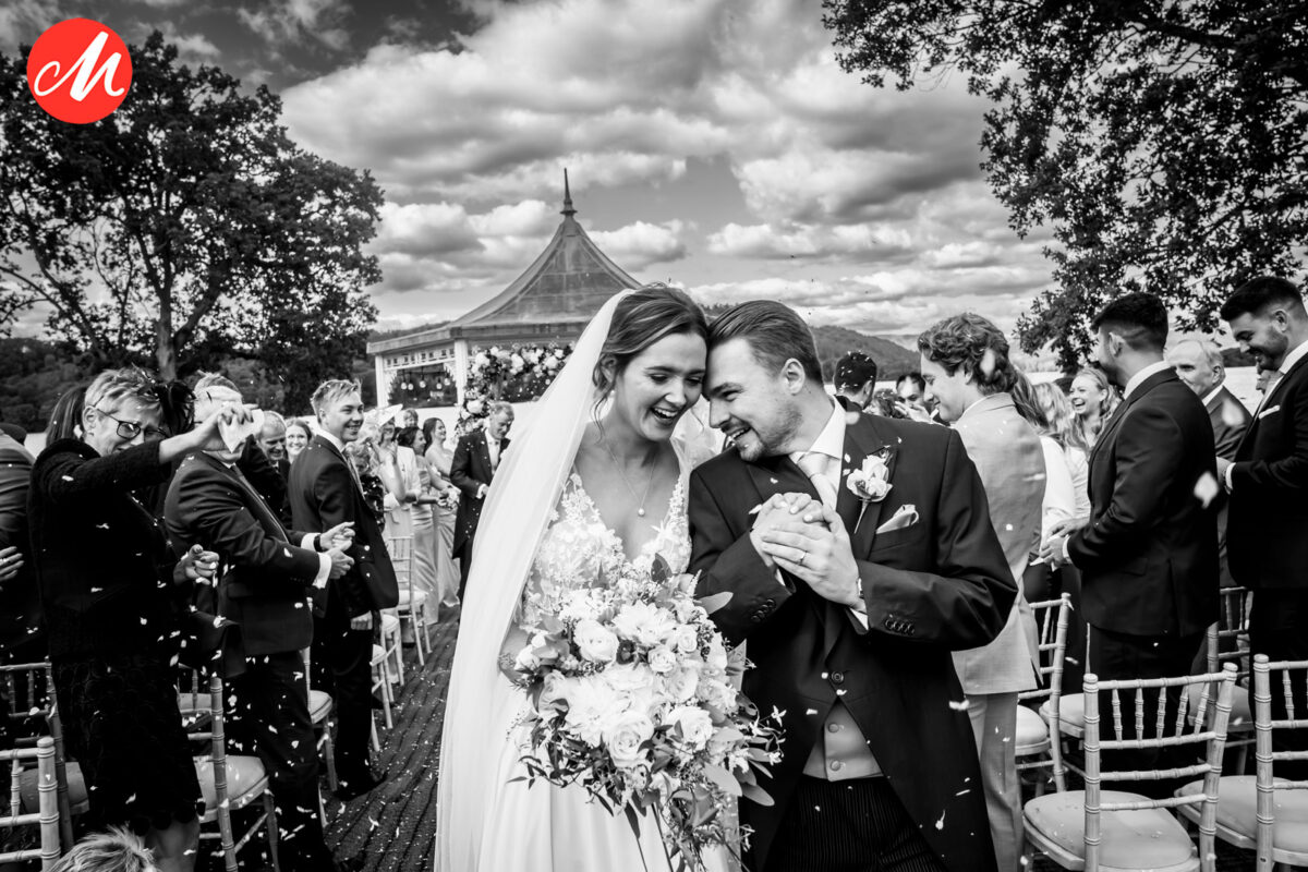 A joyous bride and groom walk down the aisle outdoors, smiling and holding hands. The bride carries a bouquet of flowers, and guests shower them with confetti. A decorative gazebo and trees are in the background under a partly cloudy sky. The scene is in black and white. Image by Andy Wade Photography.