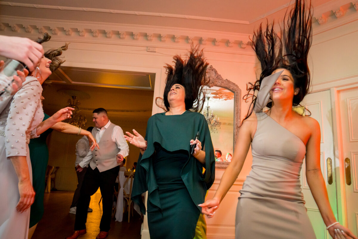 People are dancing at an indoor party; two women with dark hair are in the foreground flipping their hair up, one in a green dress and the other in a one-shoulder beige dress. The lively scene, perfect for any event portfolio, includes other guests and a lit room in the background. Image by Andy Wade Photography.