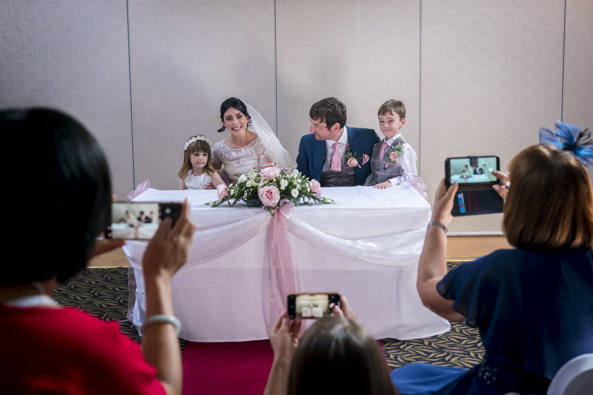A bride and groom sit at a decorated table, smiling with two children in formal attire beside them at the picturesque Stables Country Club in Lancashire. People around the table are taking photos with their phones. The table is adorned with flowers and pink ribbon, set against a plain, light-colored wall. Image by Andy Wade Photography.