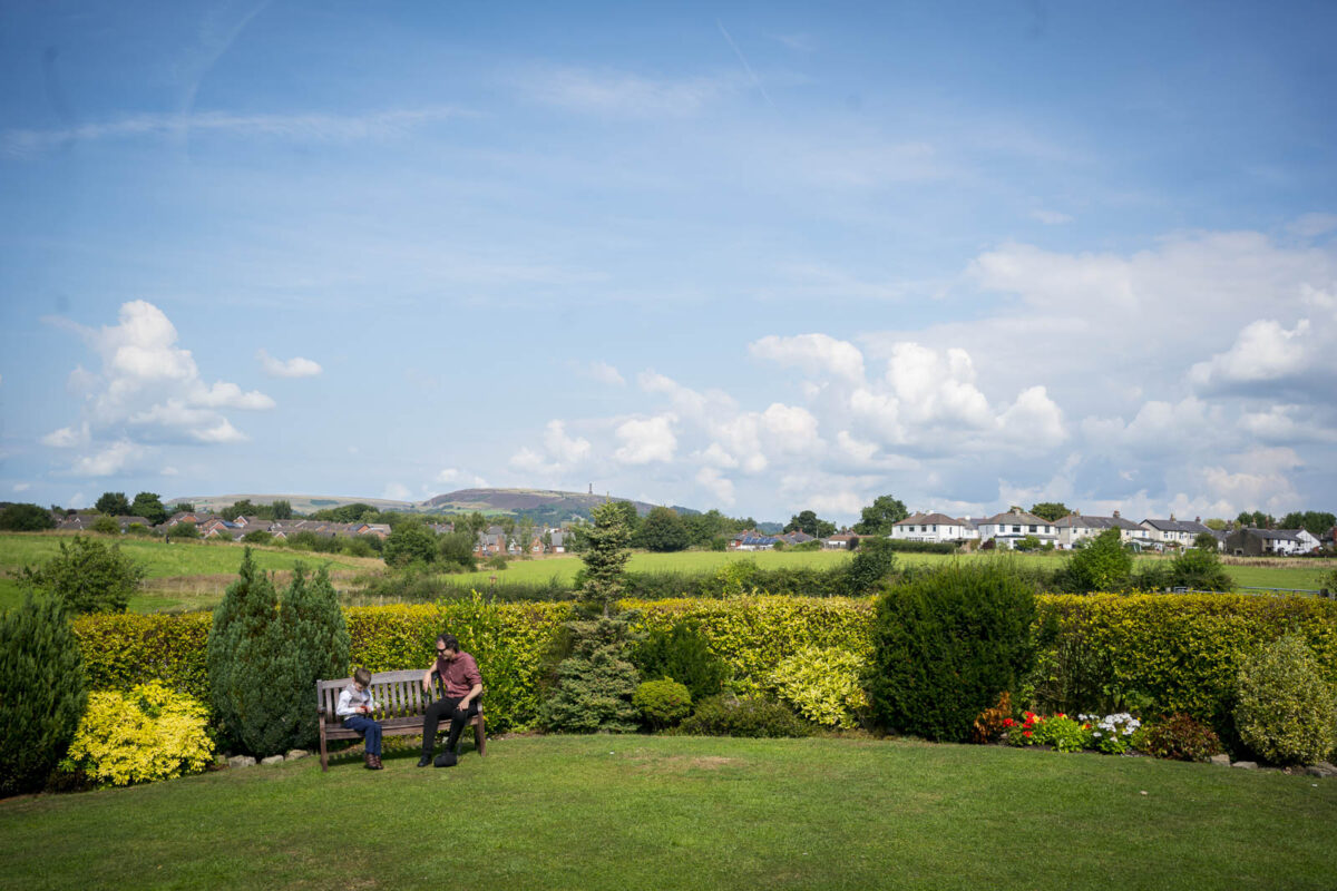 Two people sit on a bench in the garden of Stables Country Club on a sunny day, with a scenic background of rolling hills, greenery, and houses in the distance. The sky is blue with scattered clouds. The foreground has neatly trimmed grass, shrubs, and a variety of flowering plants typical of Lancashire. Image by Andy Wade Photography.