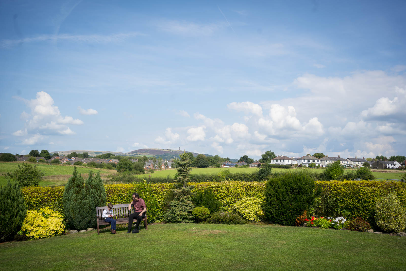 Two people sit on a bench in the garden of Stables Country Club on a sunny day, with a scenic background of rolling hills, greenery, and houses in the distance. The sky is blue with scattered clouds. The foreground has neatly trimmed grass, shrubs, and a variety of flowering plants typical of Lancashire. Image by Andy Wade Photography.