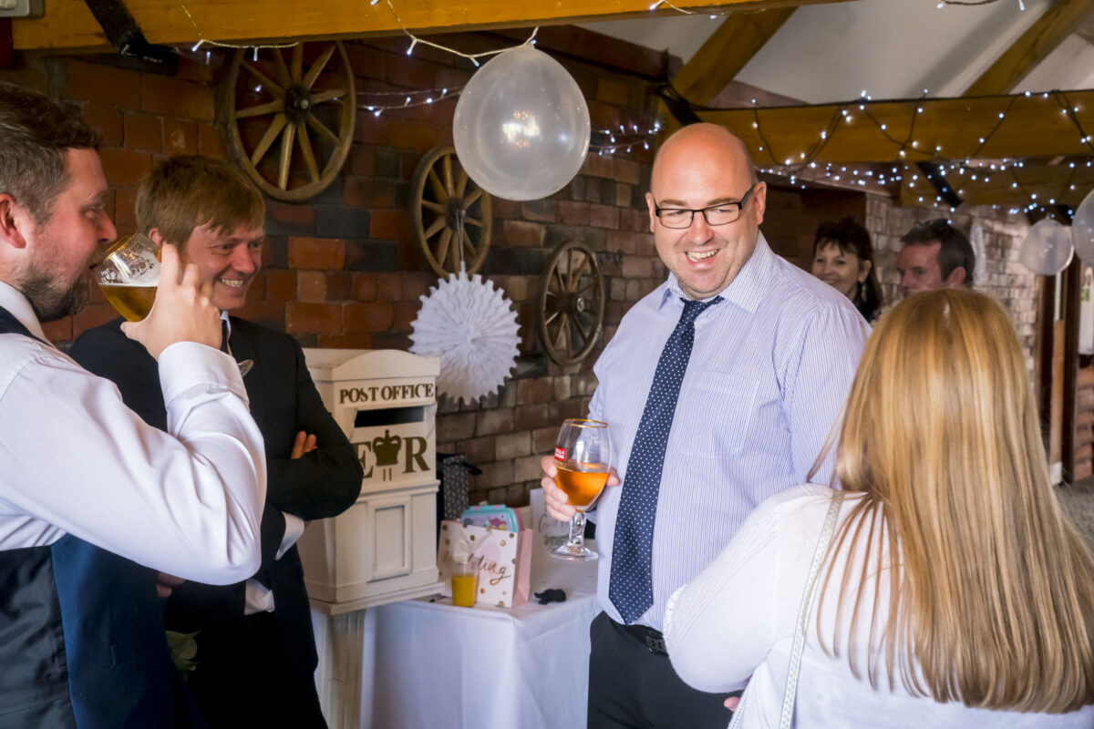 A group of four people is gathered at an indoor event near a decorated table at Stables Country Club in Lancashire. One man, wearing a suit and tie, is smiling and holding a glass. The atmosphere is festive with string lights, balloons, and a rustic brick backdrop featuring wagon wheels. Image by Andy Wade Photography.