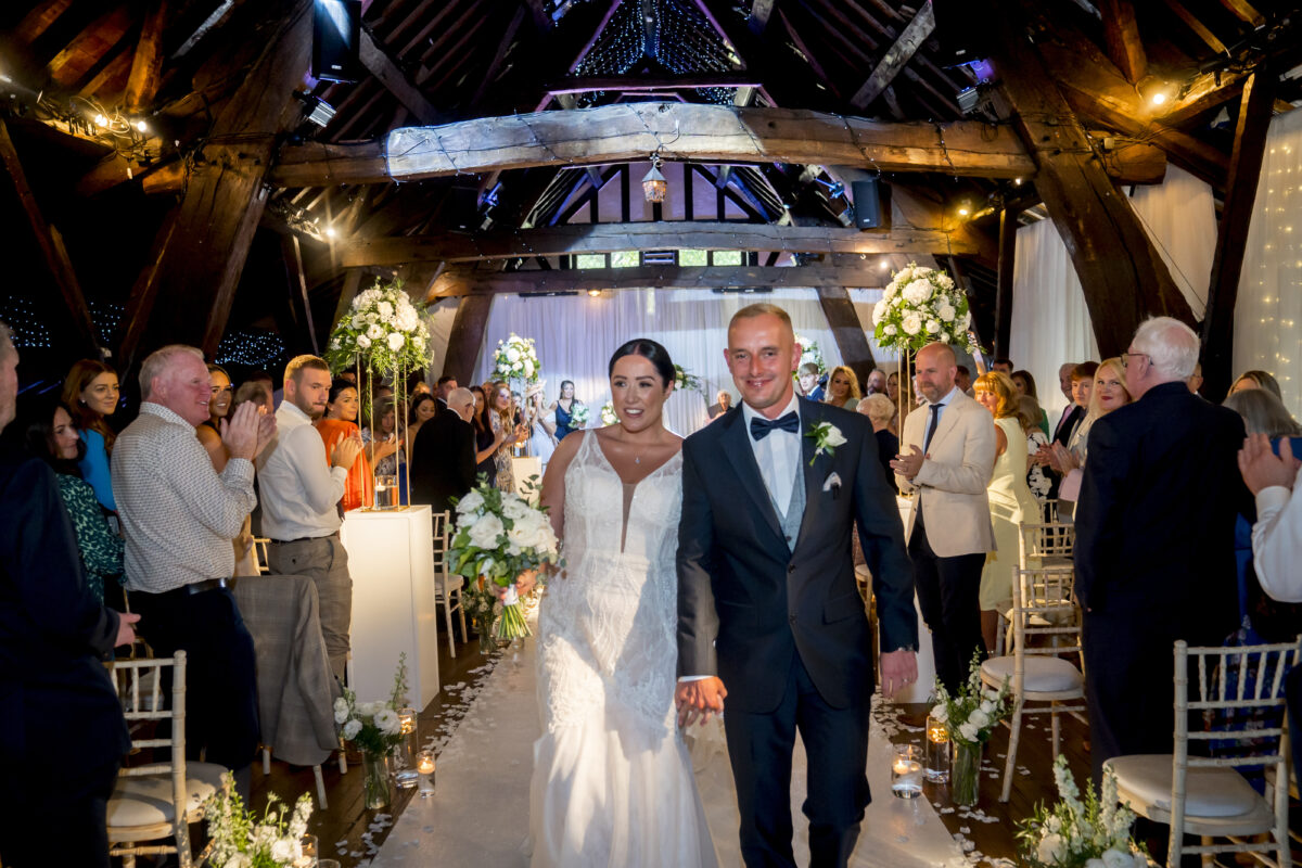 A bride in a white dress and a groom in a black suit with a bow tie walk down the aisle holding hands. Guests stand and clap on either side, with the wooden beams of Rivington Hall Barn in Lancashire visible above and floral arrangements lining the aisle. Image by Andy Wade Photography.