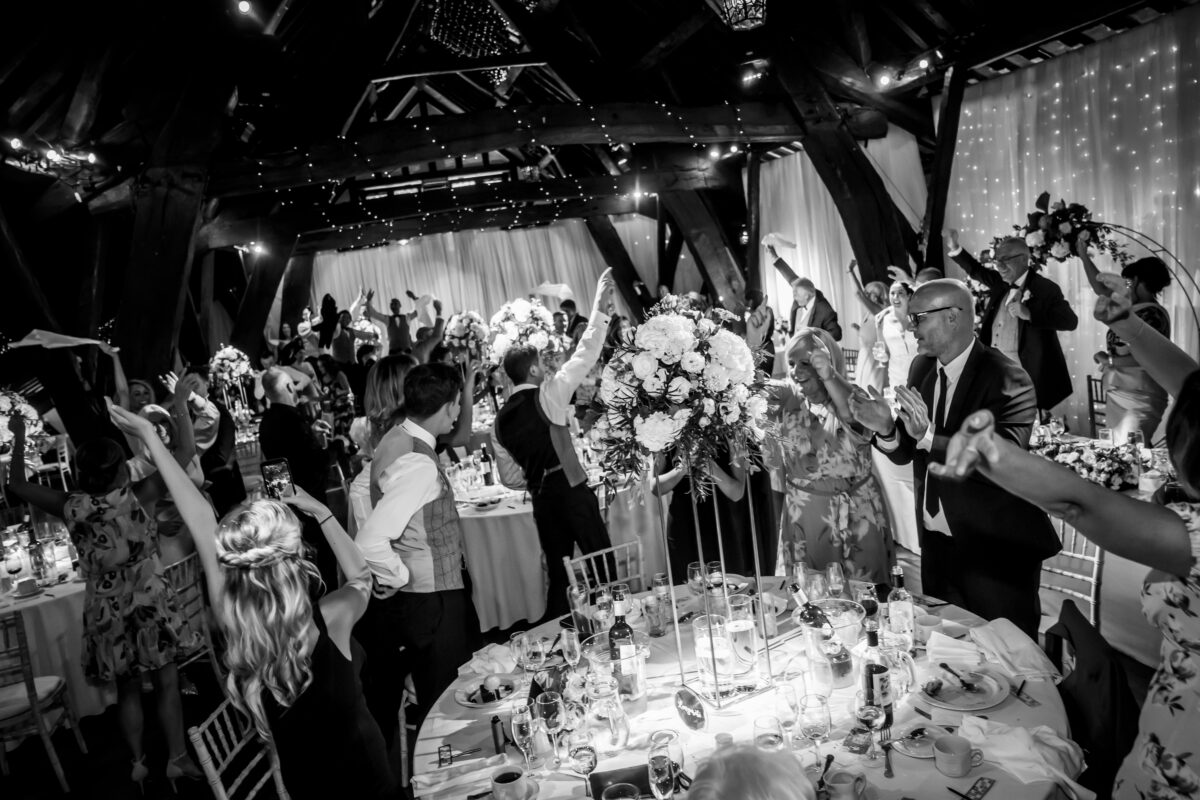 A black and white photo of a lively wedding reception at Rivington Hall Barn in Lancashire, with guests standing around tables, raising their arms in celebration. The venue features rustic wooden beams and string lights, creating a festive atmosphere. The tables are adorned with floral centerpieces and party decorations. Image by Andy Wade Photography.