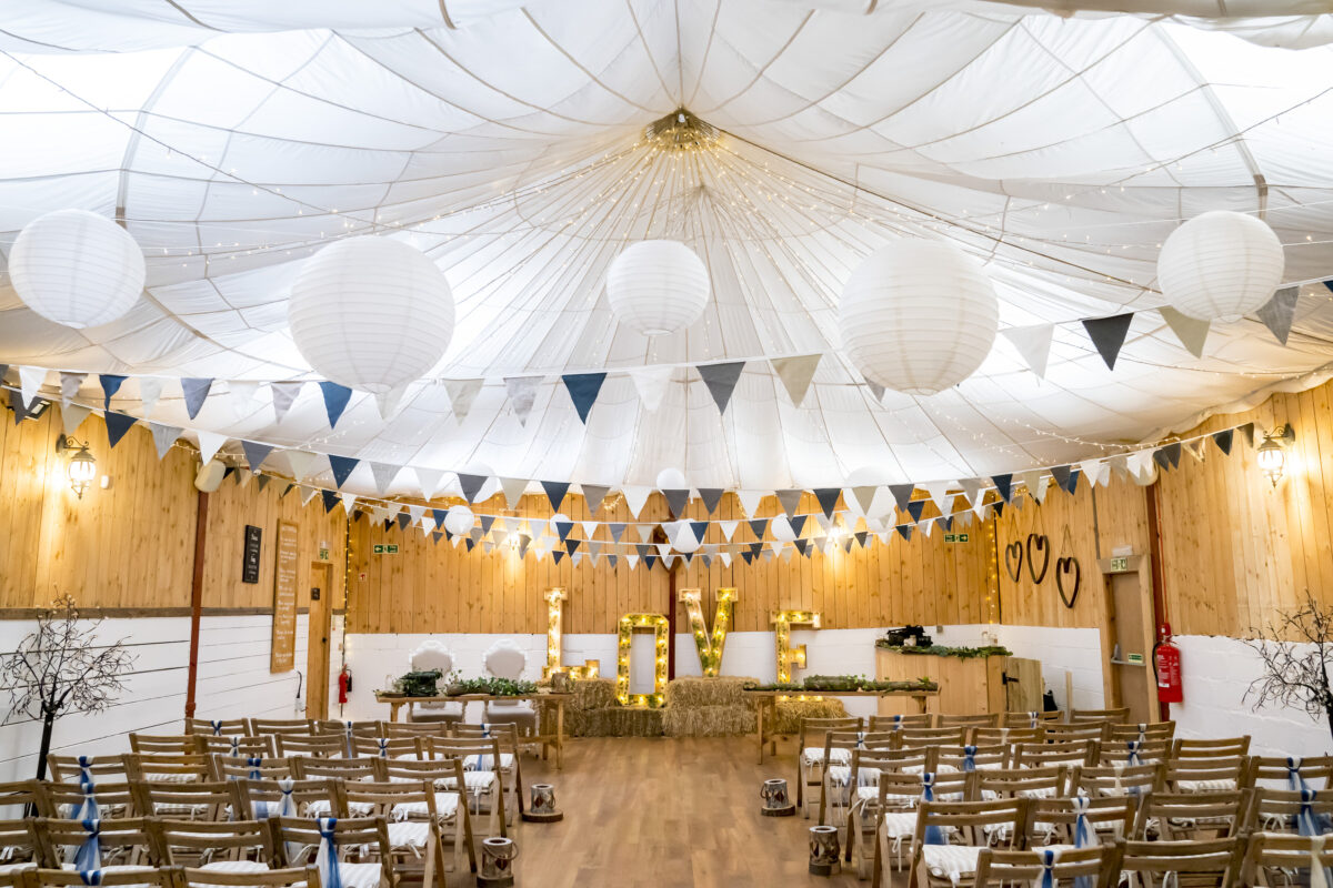 A decorated indoor wedding venue with rows of wooden chairs facing a stage. The ceiling is draped with white fabric and adorned with white hanging lanterns and string lights. A banner of white and blue pennants hangs, and large illuminated letters spelling "LOVE" sit on the stage. Captured beautifully by Andy Wade Photography at The Wellbeing Farm in Lancashire. Image by Andy Wade Photography.