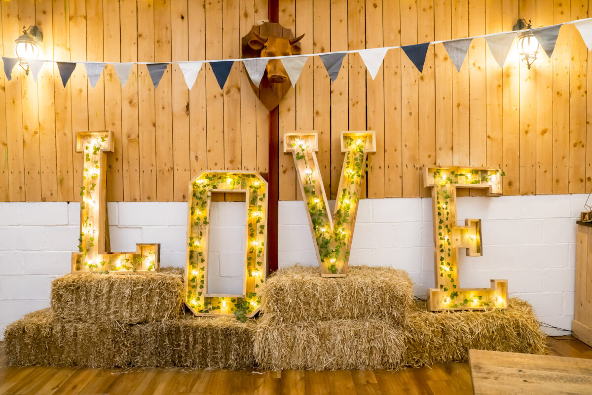 Rustic wedding decor with large illuminated letters spelling "LOVE" placed on stacks of hay bales at The Wellbeing Farm. The background features paneled wood walls, bunting in black, white, and blue colors, and wall-mounted lanterns providing warm lighting. Captured beautifully by Andy Wade Photography in Lancashire. Image by Andy Wade Photography.