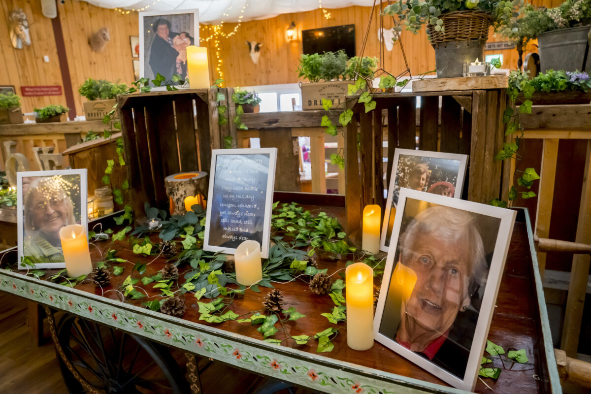 A memorial display by Andy Wade Photography features framed photographs of an elderly woman on a rustic wooden table, adorned with ivy, pine cones, and glowing LED candles. The setup is inside the cozy Lancashire venue, The Wellbeing Farm, with soft string lights in the background. Image by Andy Wade Photography.