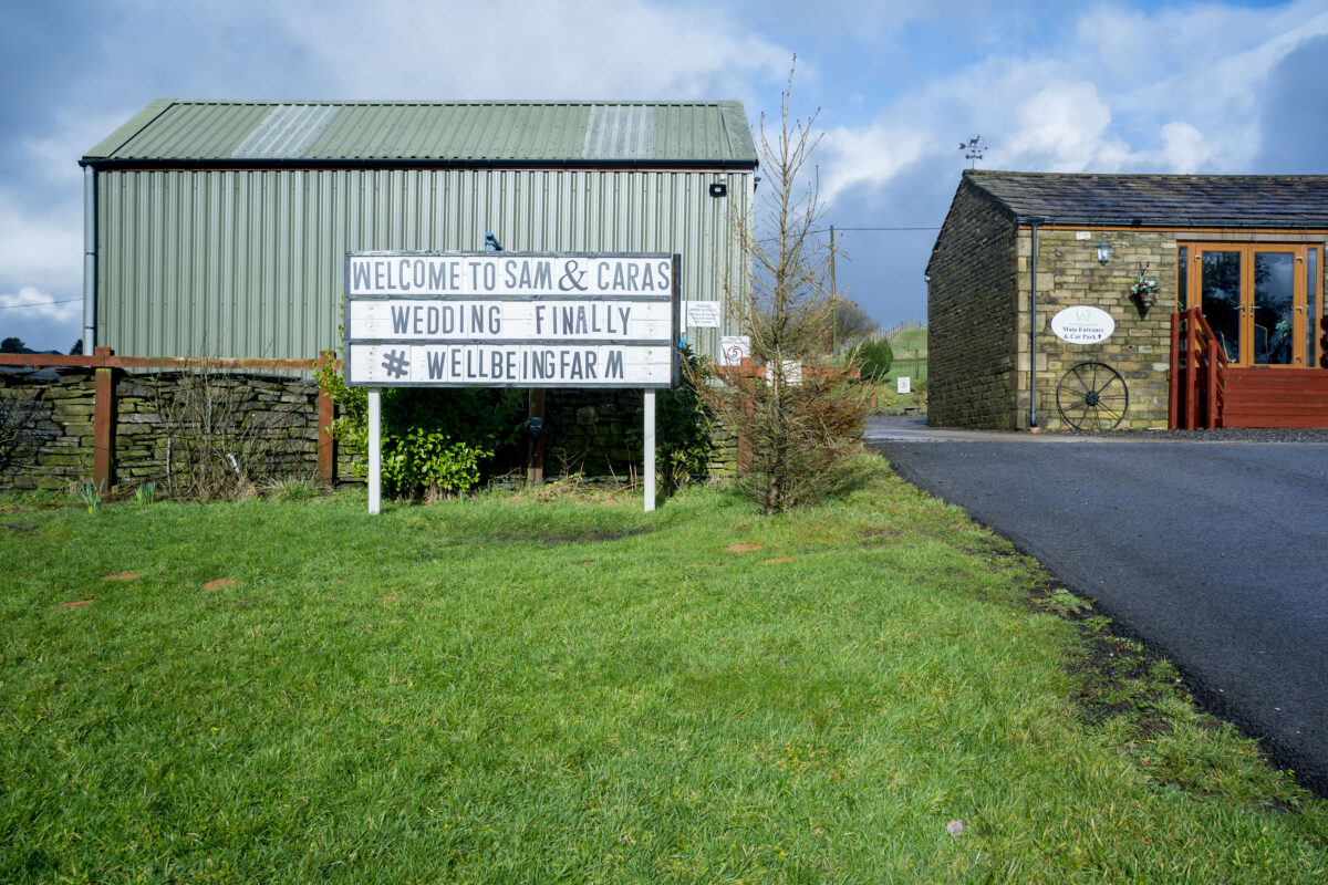 Outdoor event venue with a green lawn. A white signboard reads, "Welcome to Sam & Cara's Wedding Finally," and includes the hashtag “#wellbeingfarm.” To the right is a stone building with a wooden entrance and logo. The charming Lancashire sky is partially cloudy, captured perfectly by Andy Wade Photography. Image by Andy Wade Photography.