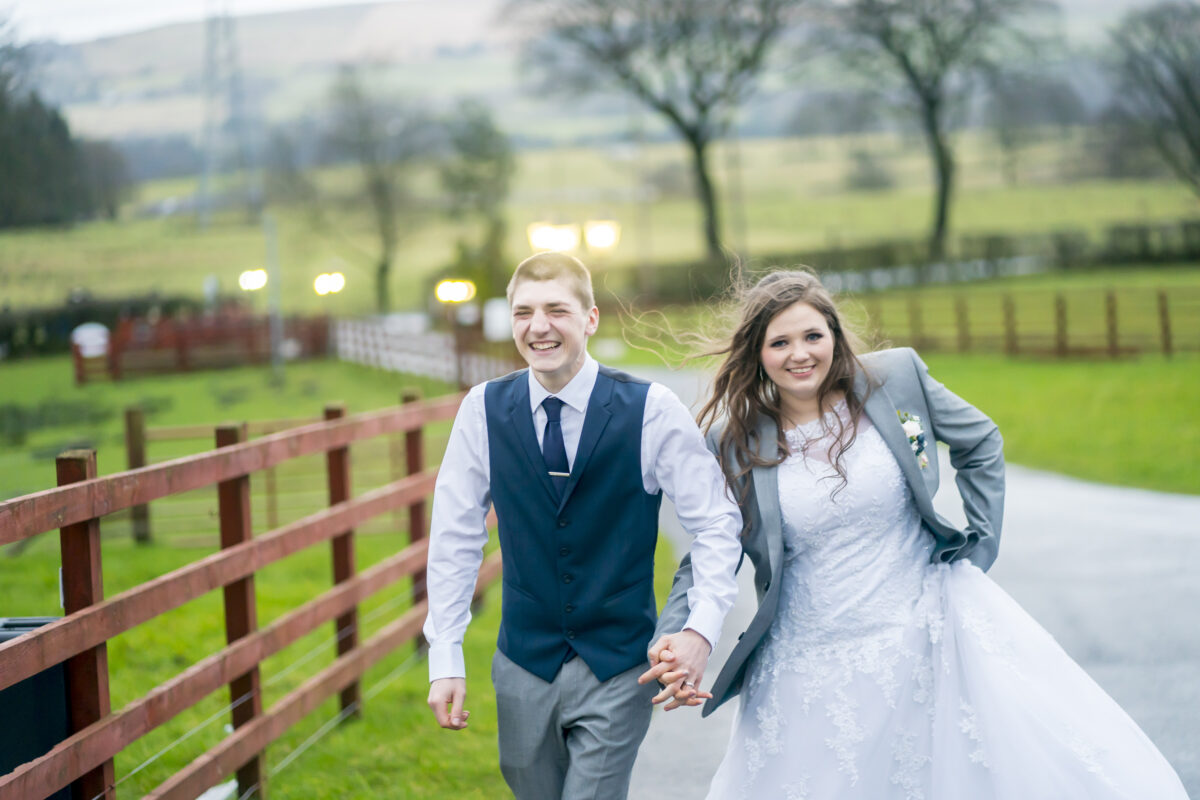 A young couple, dressed in wedding attire, walks hand in hand along a country road. The groom wears a blue vest and tie, while the bride, in a white gown with a gray jacket, smiles with her hair flowing in the wind. Captured by Andy Wade Photography at The Wellbeing Farm in Lancashire, trees and fields provide a scenic backdrop. Image by Andy Wade Photography.