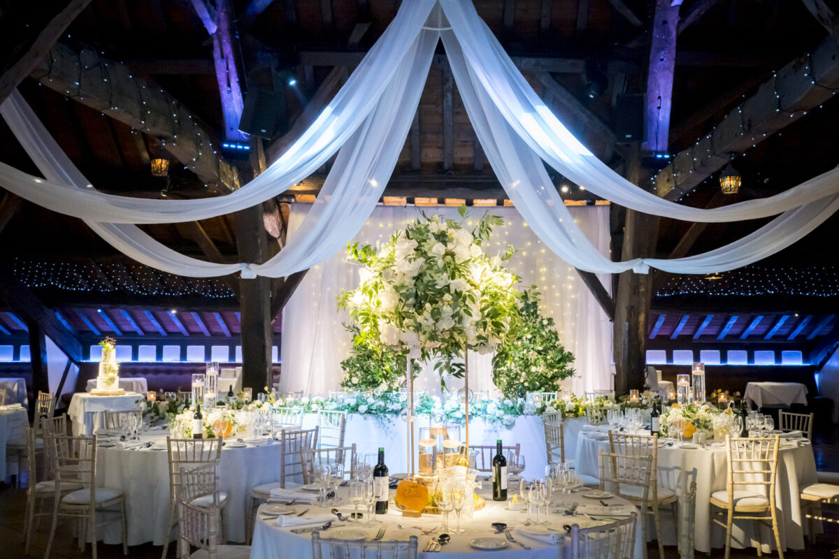 A beautifully decorated wedding reception hall in Rivington Hall Barn, Lancashire, with draped white fabric on the ceiling, elegant tables set with white chairs, floral centerpieces, and dim ambient lighting. The backdrop features green foliage and twinkling fairy lights. Image by Andy Wade Photography.