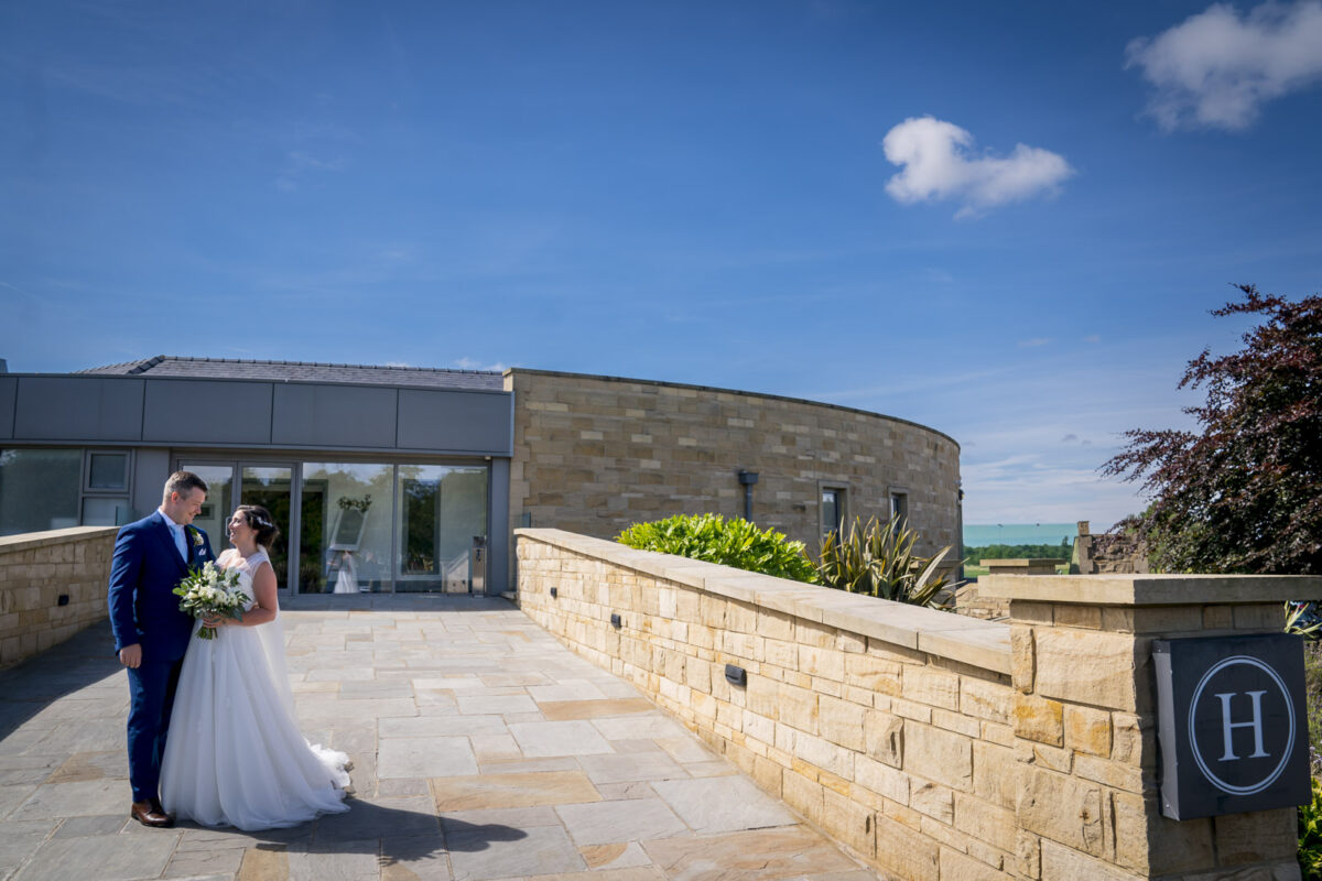 A newly married couple stands close together outdoors in front of a stone building with a curved wall at Hurlston Hall; the groom in a dark blue suit and the bride in a white gown holding a bouquet. The clear blue sky and greenery of Lancashire enhance the serene atmosphere. Image by Andy Wade Photography.