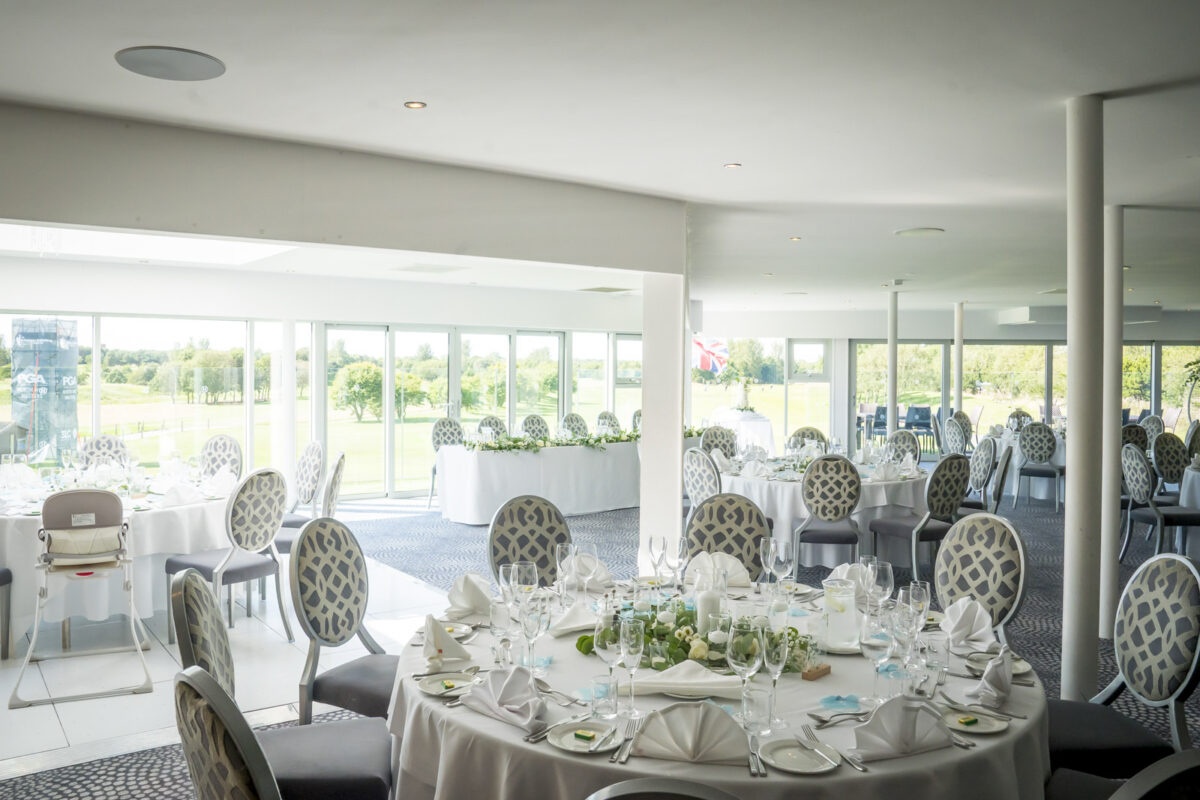 A brightly lit banquet hall at Hurlston Hall, with large windows overlooking a scenic Lancashire outdoor area. The space features round tables covered in white tablecloths, elegantly set with glassware, cutlery, and floral centerpieces. Grey upholstered chairs surround the tables. Image by Andy Wade Photography.