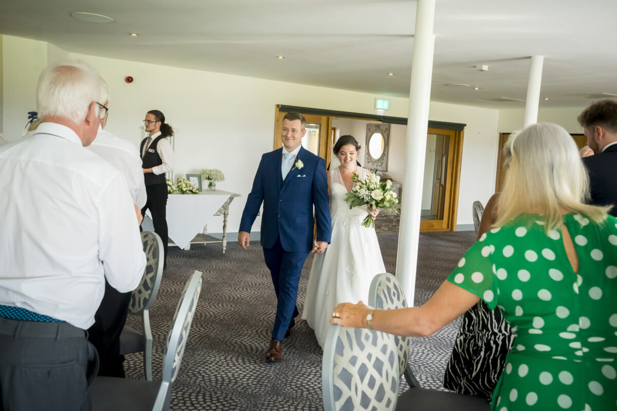 A bride in a white dress and a groom in a blue suit walk down the aisle at Hurlston Hall in Lancashire, smiling. Guests, including an older man and woman in the foreground, stand and watch. The bride holds a bouquet of white flowers while an officiant stands in the background. Image by Andy Wade Photography.