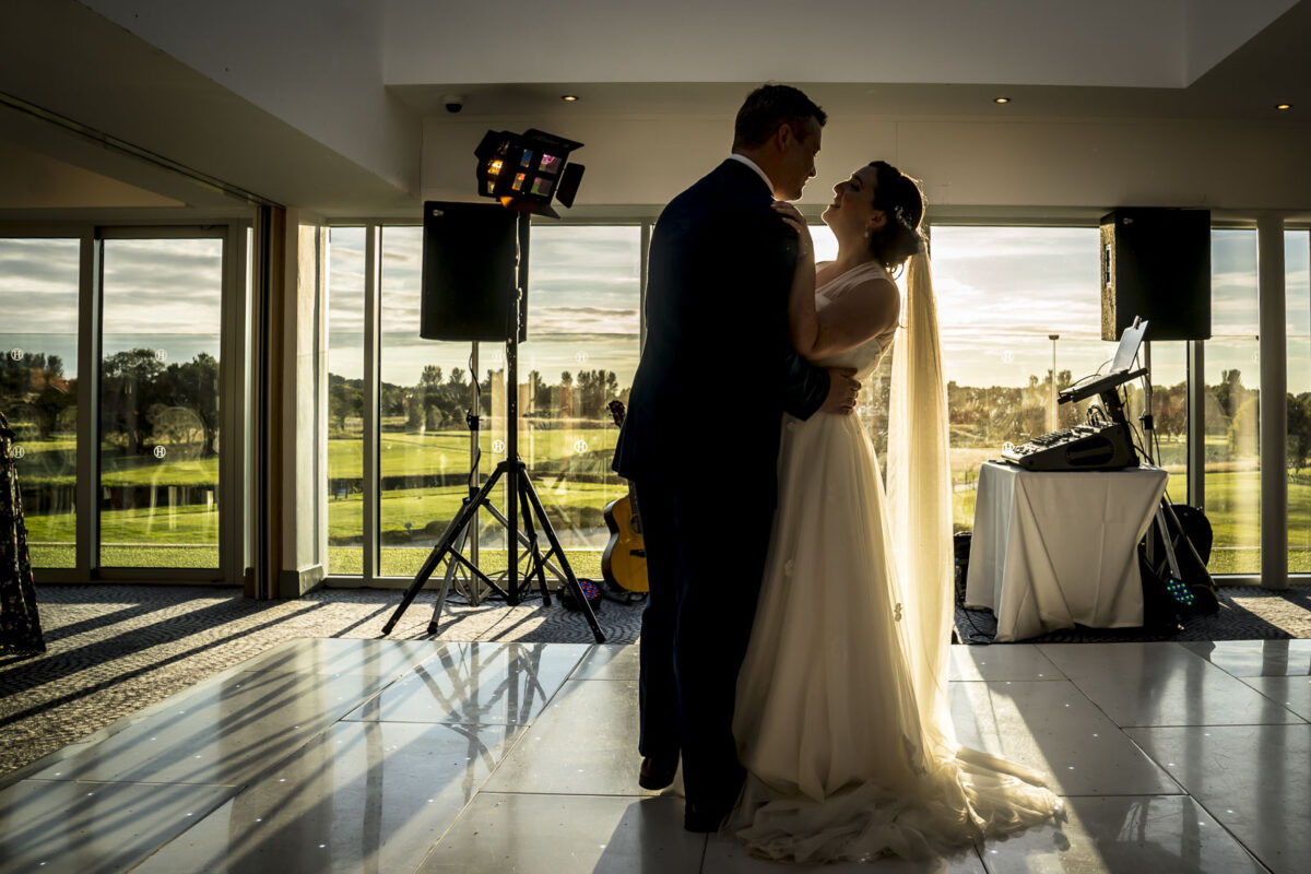 A bride and groom dance closely in a warmly lit room at Hurlston Hall with large windows overlooking the green Lancashire landscape. They are silhouetted against the soft sunlight streaming in, creating an intimate and romantic atmosphere. Musical equipment is set up nearby. Image by Andy Wade Photography.