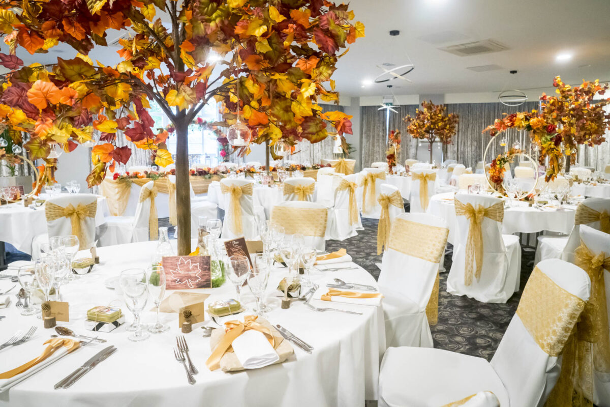 A brightly lit banquet hall at Ribby Hall is adorned with autumn-themed decor. Round tables are covered with white tablecloths, each featuring orange and gold accents, lush centerpieces with fall foliage, neatly arranged cutlery, and glassware. White chairs are tied with gold sashes. Image by Andy Wade Photography.