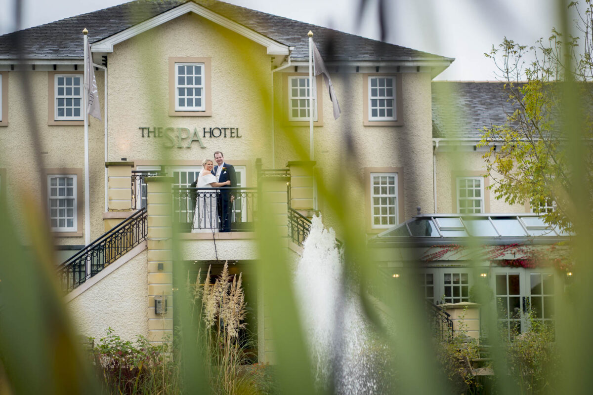 A bride and groom stand on a balcony at The Spa Hotel, smiling and embracing. The image is taken through greenery, with the hotel building as the backdrop and a water fountain visible in the foreground. This picturesque moment captures the essence of Ribby Hall in Lancashire. Image by Andy Wade Photography.
