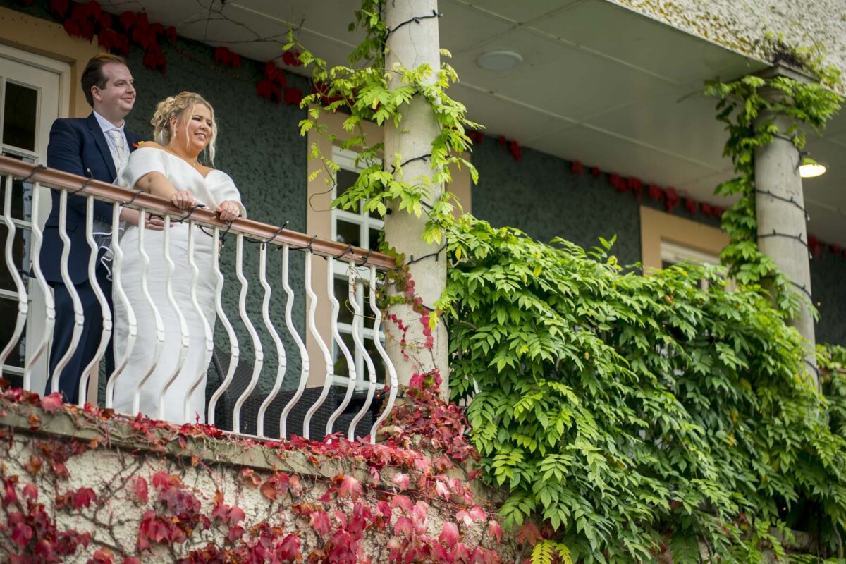 A bride and groom stand on a leafy balcony adorned with green and red ivy at Ribby Hall in Lancashire, smiling and looking out into the distance. The groom is in a navy suit, and the bride is in a white dress. The building behind them has grey and yellow accents. Image by Andy Wade Photography.