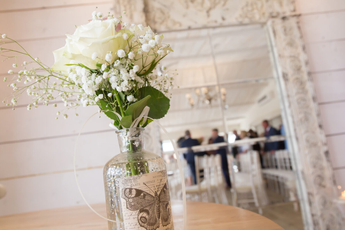 A delicate bouquet of white roses and baby's breath arranged in a vintage glass bottle sits on a wooden table. The background features a large ornate mirror reflecting a lively group at Bashall Barn, Lancashire. White chairs are arranged neatly for an elegant event. Image by Andy Wade Photography.