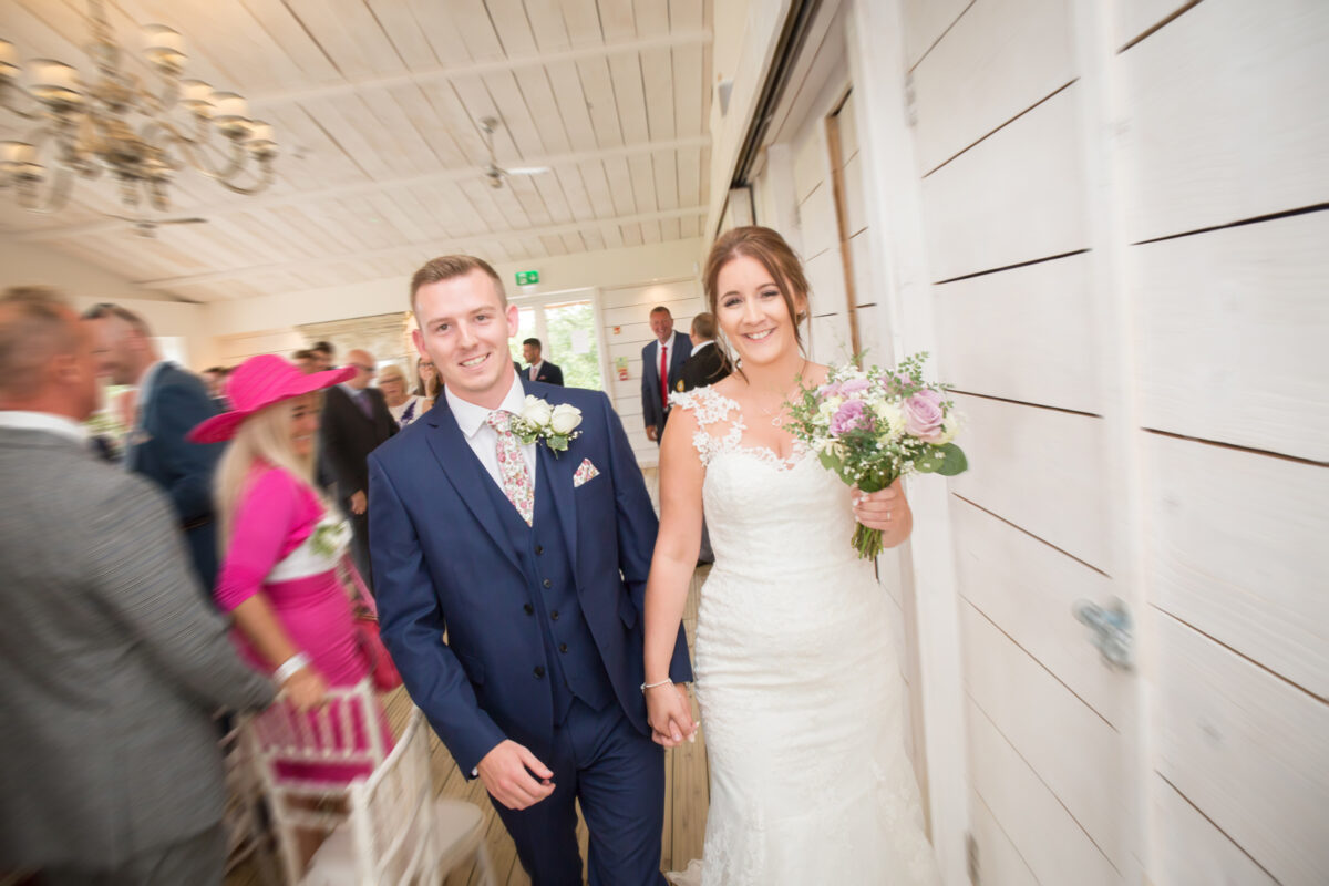 A smiling bride and groom walk hand in hand down the aisle at Bashall Barn. The bride wears a white lace wedding dress and holds a bouquet of flowers, while the groom is in a navy blue suit with a floral tie. They are in a bright room with guests clapping, some in formal attire, celebrating in Lancashire. Image by Andy Wade Photography.
