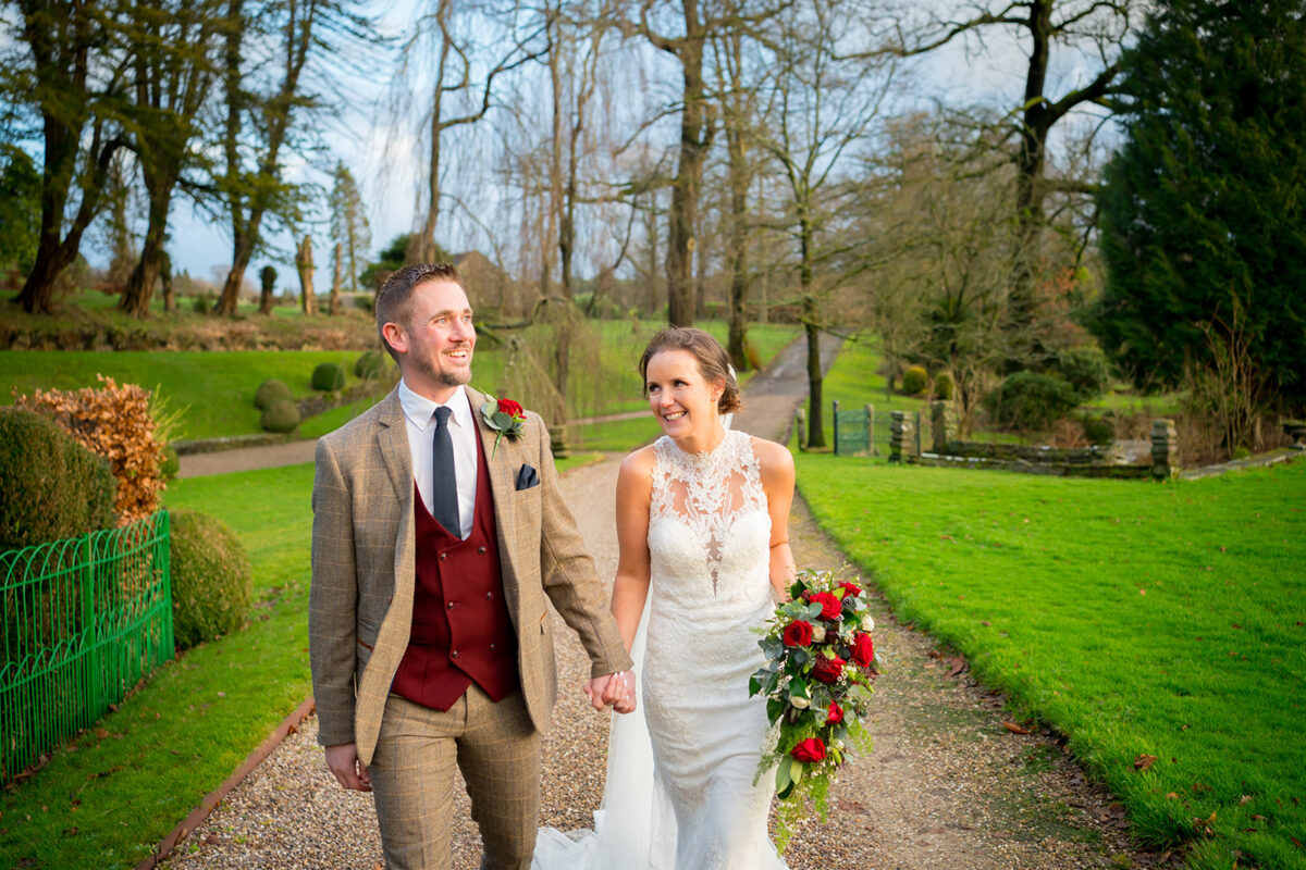 A bride and groom, dressed in elegant wedding attire, walk hand in hand along a garden path at Browsholme Hall and the Tithe Barn in Lancashire. The bride carries a bouquet of red and white flowers, while the groom sports a brown suit with a burgundy vest. The background features lush greenery and bare trees. Image by Andy Wade Photography.