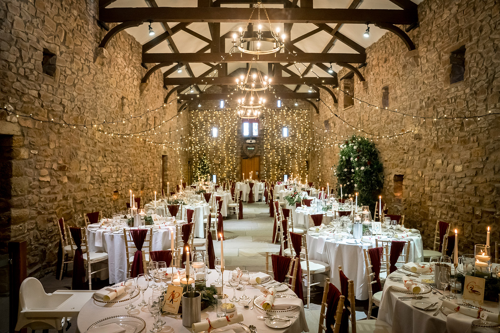A beautifully decorated banquet hall at Browsholme Hall and the Tithe Barn in Lancashire features stone walls, wooden beams, and numerous round tables set for a formal dinner. Tables are adorned with white linens, candles, and elegant place settings. Fairy lights and chandeliers provide soft, warm lighting throughout. Image by Andy Wade Photography.