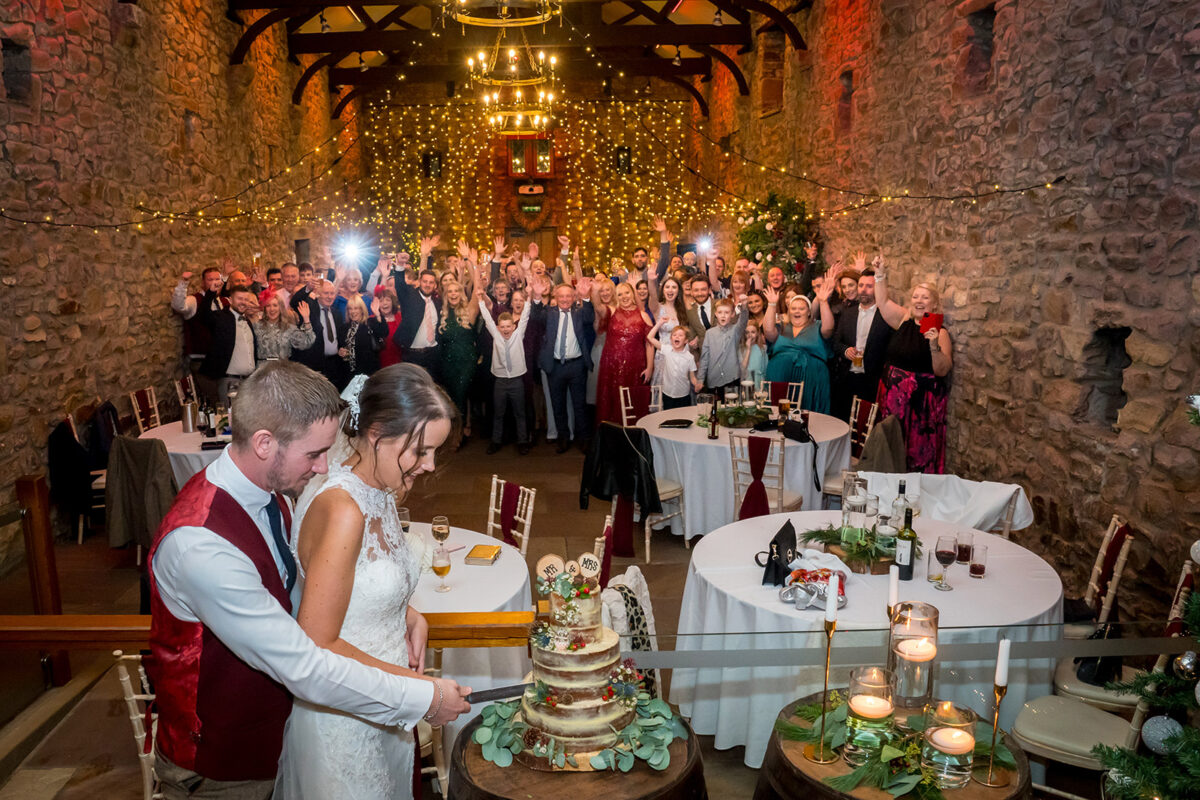 A bride and groom cut their wedding cake in the rustic Browsholme Hall and the Tithe Barn in Lancashire, surrounded by family and friends who are all cheering and raising their hands. The stone-walled room is adorned with fairy lights and chandeliers, creating a warm and festive atmosphere. Image by Andy Wade Photography.