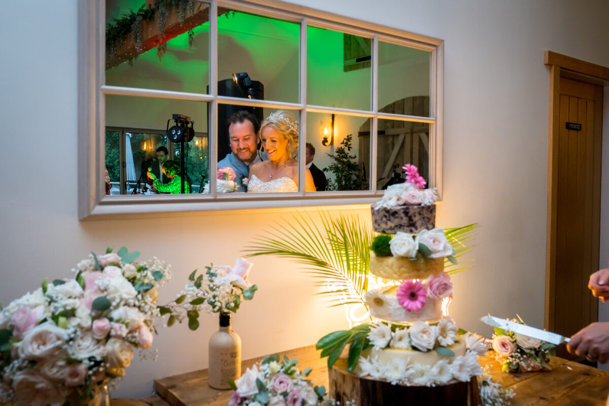 A newlywed couple is reflected in a large mirror, smiling joyfully. In the foreground, a multi-tiered wedding cake adorned with flowers is being cut. The table, set at Hobbit Hill in Lancashire, is decorated with floral arrangements and warm lighting enhances the celebratory atmosphere. Image by Andy Wade Photography.