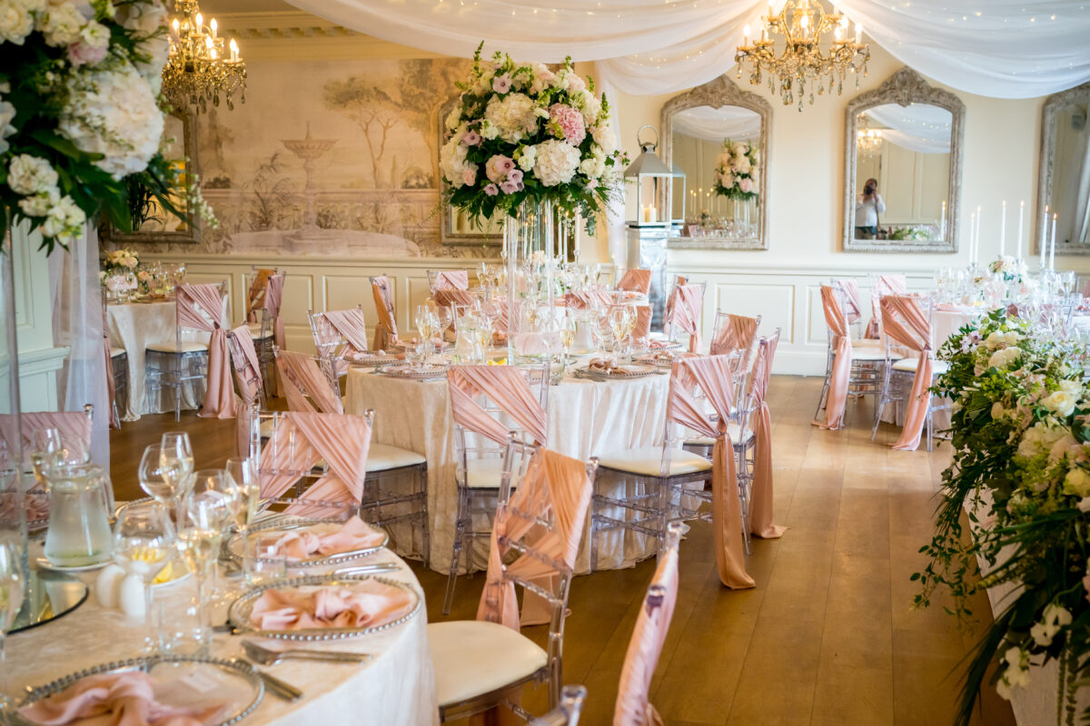 A lavish wedding reception setup at Eaves Hall in Lancashire, featuring elegantly decorated round tables covered in white tablecloths and adorned with pink chair sashes. The room boasts floral centerpieces, a white draped ceiling, chandeliers, and large mirrors on the walls, creating a sophisticated ambiance. Image by Andy Wade Photography.