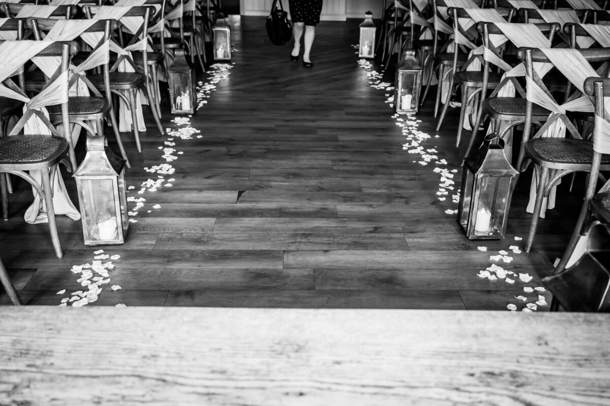 Black and white image of an empty wedding aisle decorated with lanterns and flower petals at the Shireburn Arms in Lancashire. Cross-back wooden chairs line both sides of the aisle. A person is walking away from the camera at the end of the aisle. The floor is wooden. Image by Andy Wade Photography.