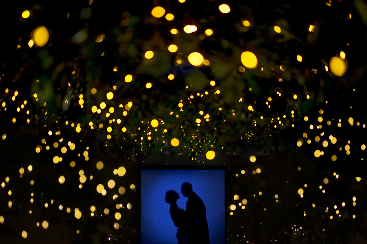 A silhouetted couple embraces against a vibrant blue backdrop, framed by a multitude of glowing yellow lights that create a dreamy, star-like effect surrounding them in the darkness at Stirk House in Lancashire. Image by Andy Wade Photography.