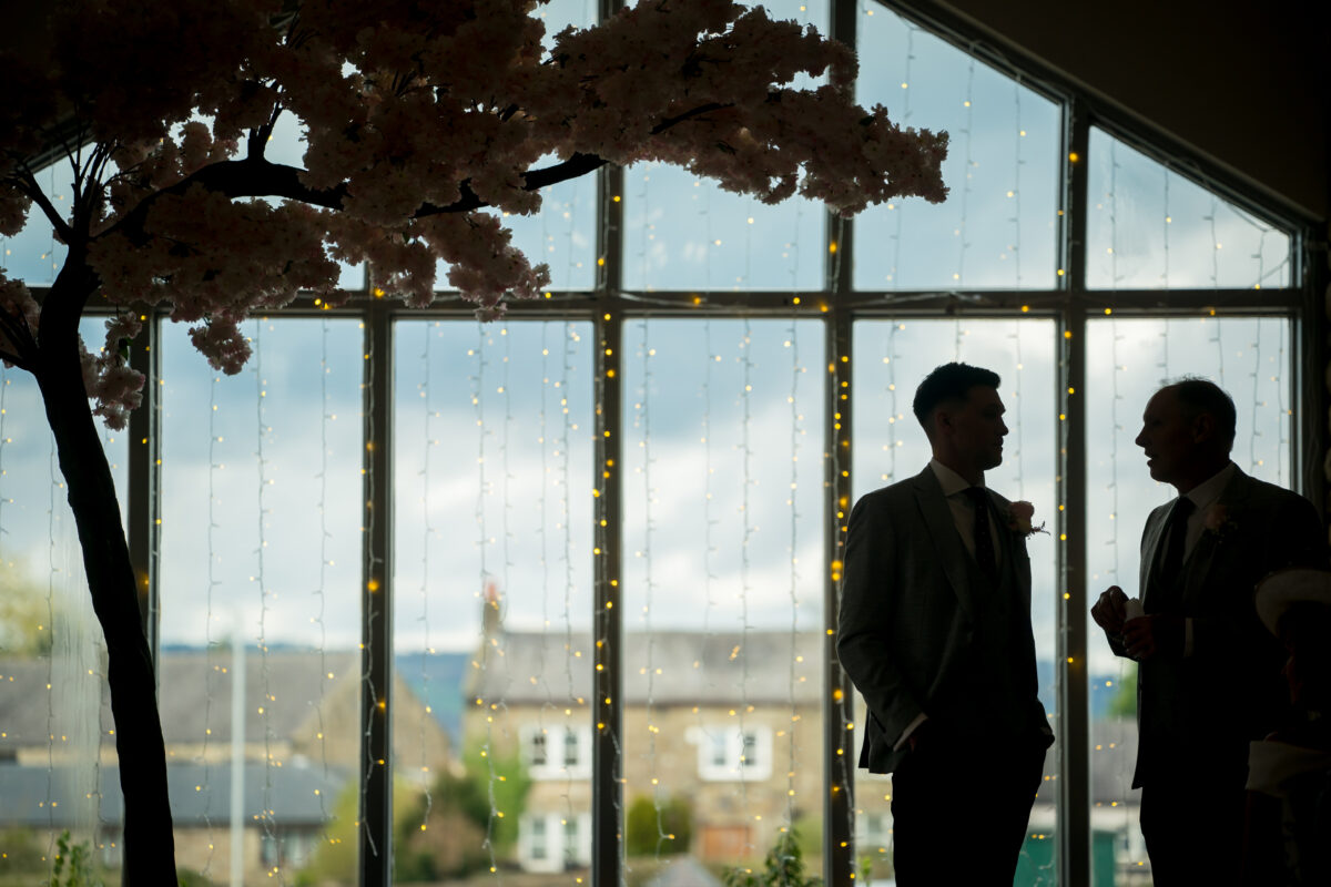 Two individuals in formal attire are silhouetted against a large window with twinkling string lights at Mytton Fold. A decorative tree branch hangs overhead, adding to the serene and intimate ambiance. The view outside reveals a cloudy Lancashire sky and distant buildings. Image by Andy Wade Photography.