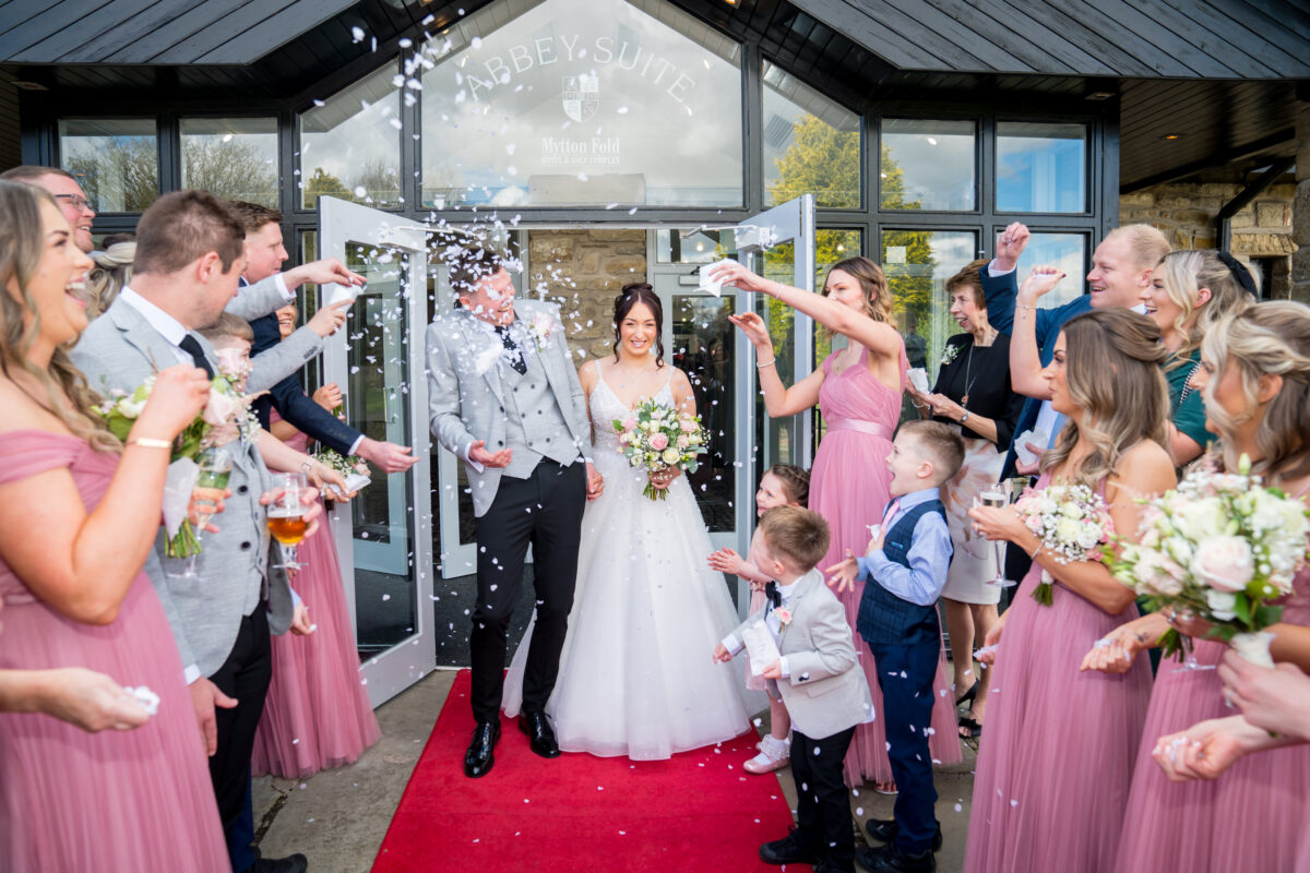 A newlywed couple exits Mytton Fold in Lancashire as guests throw confetti. The bride, in an elegant white gown, holds a bouquet, while the groom, in a grey suit, smiles beside her. Bridesmaids in pink dresses and other guests, including children, cheer around them on a red carpet. Image by Andy Wade Photography.
