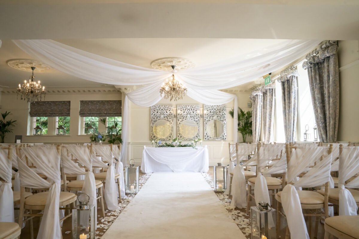 A beautifully decorated wedding ceremony venue at Eaves Hall in Lancashire, featuring white chairs draped in white fabric and adorned with flowers, arranged in rows along a white aisle covered in petals. Elegant chandeliers hang from the ceiling, and mirrors with intricate frames are displayed at the front. Image by Andy Wade Photography.