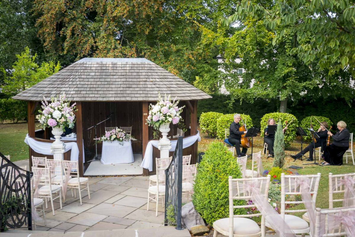 A wooden gazebo adorned with floral arrangements is set up for a wedding ceremony at Ribby Hall in Lancashire. White chairs are arranged in rows facing the gazebo. A string quartet is seated to the right, preparing to play. The area is surrounded by lush greenery and trees. Image by Andy Wade Photography.