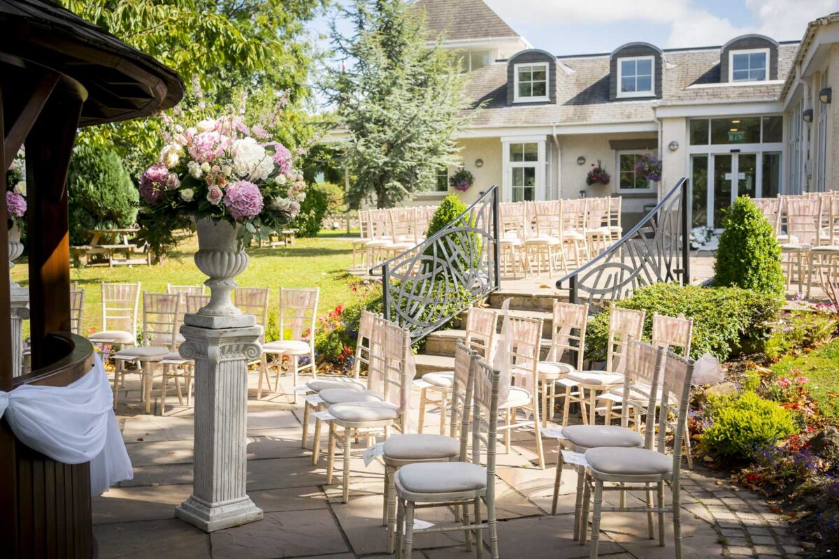 Outdoor wedding setup at Ribby Hall in Lancashire featuring rows of white chairs arranged before a grand building with large windows. A stone pedestal with a bouquet of pink and white flowers stands nearby. Pathways, greenery, and a staircase leading to a garden enhance the elegant setting. Image by Andy Wade Photography.