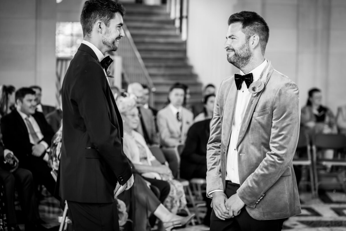 A black and white image shows two men, both dressed formally in suits, smiling and making eye contact at a wedding ceremony in Lancashire. They stand in front of seated guests who appear to be watching them with interest. A staircase is visible in the background, possibly at The Mill at Condor Green. Image by Andy Wade Photography.