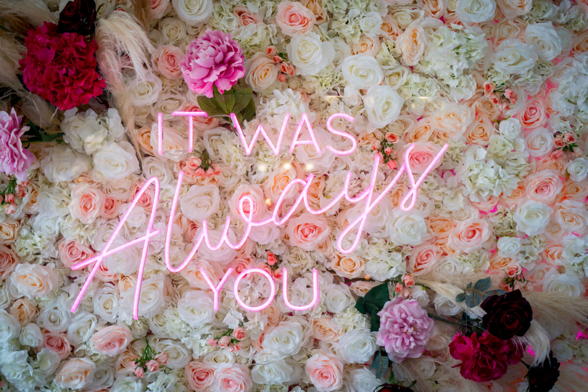 A neon sign displaying the message "IT WAS Always YOU" is mounted on a vibrant floral wall at The Mill at Condor Green. The wall features a variety of colorful flowers, including roses, peonies, and hydrangeas in shades of pink, white, and red, creating a lush and romantic background perfect for any Lancashire setting. Image by Andy Wade Photography.