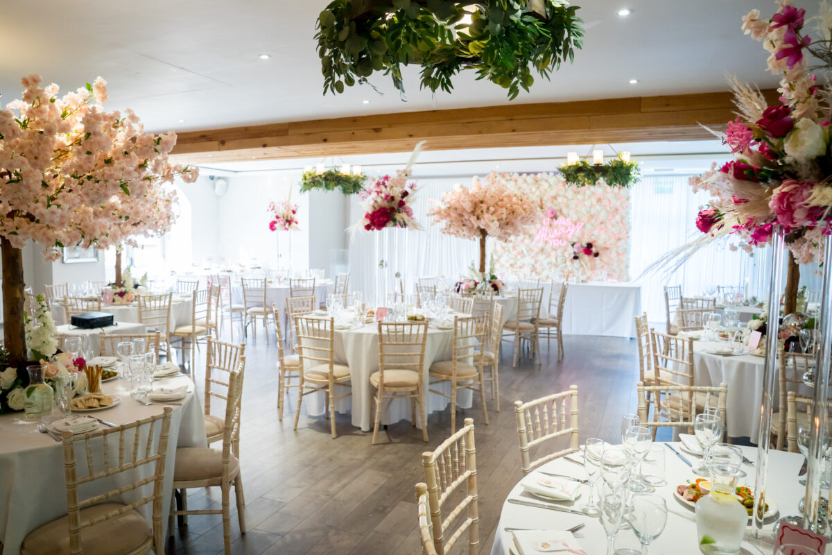 A spacious and elegant event hall at The Mill at Condor Green, Lancashire, decorated for a wedding reception. The room features round tables set with white linens, glassware, and floral centerpieces. The ceiling and tables are adorned with an abundance of lush pink and white blossom arrangements. Image by Andy Wade Photography.