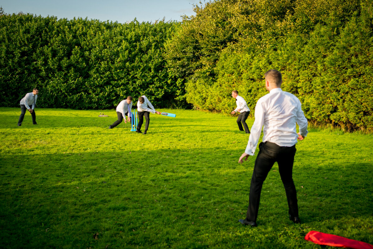 Five people are playing cricket on a grassy field near The Mill at Condor Green, Lancashire. The batter is about to hit the ball with a blue bat while the other four participants are positioned around the field. All are dressed in white shirts and black pants, with greenery and trees in the background. Image by Andy Wade Photography.